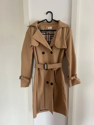 Beige trenchcoat med bälte - Stilren beige trenchcoat från JIEPOTU med klassiskt rutigt foder, dubbelknäppning och bälte i midjan. Jackan har bred krage, axelklaffar och justerbara spännen vid ärmsluten. Perfekt för dig som vill ha en tidlös och trendig look. ALDRIG ANVÄND