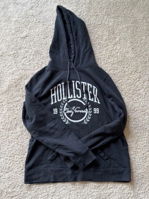 Svart hoodie från Hollister - Svart hoodie från Hollister med stor vit logga och texten 'California 1999' på bröstet. Tröjan har huva med dragsko och känguruficka framtill. Mjuk insida i fleece och ribbade muddar vid ärmslut och nederkant. Perfekt för en avslappnad och cool stil.