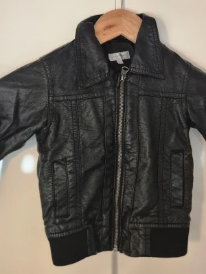 Svart skinnjacka med dragkedja - Snygg svart skinnjacka med klassisk krage och silverfärgad dragkedja framtill. Jackan har två fickor på sidorna och ribbade muddar vid ärmslut och nederkant för en cool look. Perfekt för dig som vill ha en tidlös och stilren jacka.i storlek 80