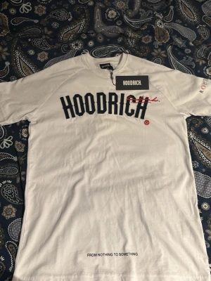 Vit HOODRICH t-shirt med tryck XS - Vit t-shirt från HOODRICH med svart och rött tryck framtill. Klassisk rund hals och korta ärmar. Texten 'FROM NOTHING TO SOMETHING' finns tryckt längst ner. Perfekt för en streetwear-look.