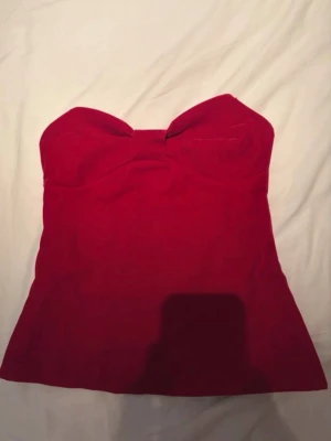 Röd bandeau topp med rosett - Supersnygg röd bandeau topp med en stor rosett framtill. Toppen är axelbandslös och har en figurnära passform som framhäver siluetten. Perfekt för dig som vill sticka ut lite extra med en söt detalj.