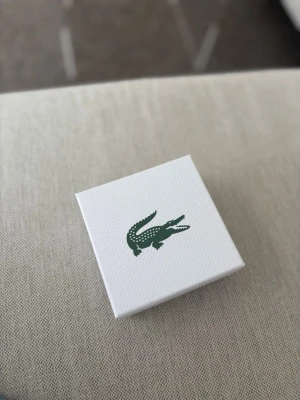 Silverfärgat armband från Lacoste - Snyggt armband i silverfärgad metall från Lacoste. Armbandet har en modern, fyrkantig länkdesign med graverade detaljer och den klassiska krokodilloggan. Perfekt accessoar för dig som gillar stilrena och sportiga smycken.