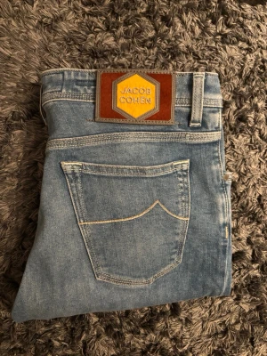 Jacob Cohen - Säljer ett par ljusblå jeans från Jacob Cohen med klassisk femficksmodell och snygga kontrastsömmar. Jeansen har en patch i brunt och gult läder bak i midjan och är tillverkade i mjukt denimtyg. Perfekta för en avslappnad och trendig look.