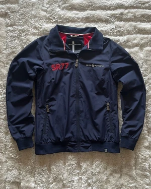 Sail Racing jacka i marinblå - stl M - Sail Racing jacka i marinblå, storlek M men sitter som en liten M. Skal 100% nylon, foder 100% polyester, dragkedja hela vägen, två fickor med dragkedja, ribbade muddar och linning. Broderad logga på bröstet och tryck på ryggen.  Använd men i mycket fint skick, inga hål eller fläckar.  Rök- och djurfritt hem. Skriv gärna om du vill ha fler bilder eller undrar något!