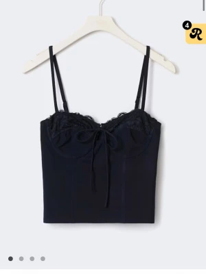 Mörk blå corset top - Trendig svart crop top med tunna axelband och spetsdetaljer vid bysten. Toppen har en söt rosett framtill och en figurnära passform som framhäver midjan. Perfekt att matcha med jeans eller kjol för en cool och stilren look.