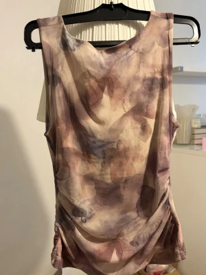 Ljuslila mönstrad ärmlös linnetopp - Säljer en ärmlös linnetopp i ljusa toner av lila, beige och rosa med ett abstrakt mönster. Toppen har rund rygg och är lätt figursydd med drapering i sidorna. Perfekt för dig som gillar en feminin och trendig stil.