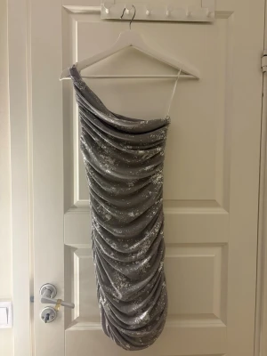 Glittrig oneshoulder silverklänning - Säljer en glittrig silverfärgad kortklänning med oneshoulder-design. Klänningen har ett smalt axelband och är täckt av glittriga detaljer. 