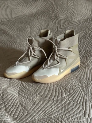 Nike Air Fear of God 1 “Oatmeal” - Säljer ett par riktigt snygga high-top sneakers från Nike i beige och vitt. Skorna har en futuristisk design med högt skaft, grova snören och en chunky sula i gummi. Materialmix av textil och syntet ger en clean och modern vibe. Nike Air Fear of God 1 “Oatmeal”. Det där är storleken:  * M10 = US herrstorlek 10 * W11.5 = US damstorlek 11,5 * Motsvarar ungefär EU 44 / 28 cm