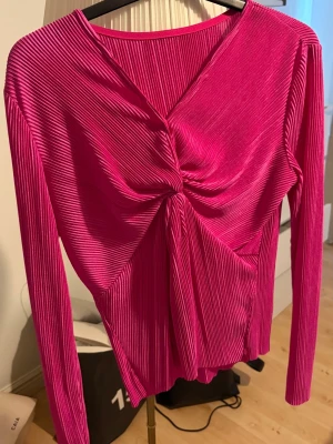 Rosa omlottopp med twist framtill - Snygg rosa topp med omlottdesign och twistad detalj framtill. Toppen har lång ärm och är gjord i ett glansigt, ribbat material som ger en cool struktur. Perfekt för dig som vill sticka ut lite extra.