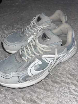 Grå b30 - Snygga sneakers i grått och vitt med meshpaneler och vita detaljer. Skorna har en sportig look med tjock sula och vita skosnören. Perfekta för dig som gillar en clean och modern stil. Märket syns på plösen och sidan.
