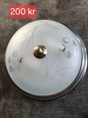 Rund taklampa i frostat glas - Säljer en rund taklampa i frostat glas med blommönster och metallkant. Lampan har en dekorativ mässingsfärgad knopp i mitten och plats för två glödlampor. Passar perfekt för dig som vill ha en stilren och klassisk belysning. Man får skaffa en kontakt.