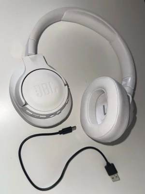 Vita over-ear hörlurar från JBL - Snygga vita over-ear hörlurar från JBL med mjuka, vadderade kåpor och justerbart huvudband. Trådlös modell med USB-C laddkabel. Modern design i plast och konstläder som ger både stil och komfort till vardagslyssningen.