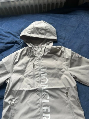 Ljusgrå vindjacka från Moncler - Snygg ljusgrå vindjacka från Moncler med huva och dragkedja framtill. Jackan har två sidofickor med dragkedja, en bröstficka och Moncler-logga på ärmen. Stora bokstäver på framsidan och ventilerande panel bak. Perfekt för blåsiga dagar.
