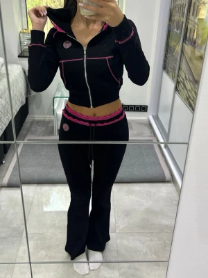 Svart loungewear set med rosa detaljer - Säljer ett svart loungewear set med dragkedja och huva på tröjan samt rosa kontrastsömmar och spetsdetaljer. Byxorna har utsvängda ben, dragsko i midjan och matchande rosa spetskant. Perfekt för chill dagar hemma.