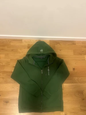 Grön hoodie från Sail Racing - Snygg grön hoodie från Sail Racing med dragkedja framtill och broderad logga på bröstet. Hoodien har en huva med tryckt logga, ribbade muddar och en bröstficka med dragkedja. Tillverkad i mjukt material som känns skönt mot huden.