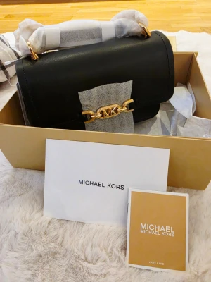 Svart handväska från Michael Kors - Snygg svart handväska från Michael Kors i slätt läder med guldfärgade detaljer och kedja. Klassisk rektangulär form med lock och MK-logga framtill. Levereras i originalkartong med tillhörande kort och dustbag.