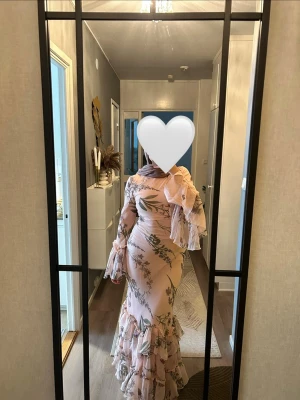 Ljusrosa långklänning med volanger - Säljer en elegant ljusrosa långklänning med blommigt mönster och volangdetaljer. Klänningen har lång ärm med volang vid ärmslut och axel, samt figurnära passform. Perfekt för dig som vill sticka ut med en romantisk look.