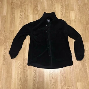 Svart overshirt i manchester från H&M - Säljer en svart overshirt från H&M i mjuk manchester. Skjortan har knäppning framtill, klassisk krage och två bröstfickor. Perfekt att bära öppen eller stängd för en avslappnad look.