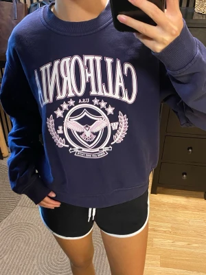 Mörkblå sweatshirt med California-tryck - Mörkblå sweatshirt med stort rosa och vitt 'CALIFORNIA' tryck på bröstet. Tröjan har rund hals, ribbade muddar och avslappnad passform. Perfekt för en chill och trendig look. Storlek M, men passar även mig som är XS/S!