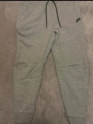 Gråa mjukisbyxor från Nike - Säljer ett par gråa mjukisbyxor från Nike med svarta dragsnören och logga på vänster sida. Byxorna har muddar vid bensluten, fickor på sidorna och är tillverkade i mjukt sweatshirtmaterial. Perfekta för chill eller träning.