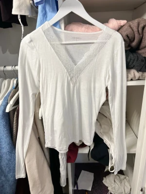 Vit långärmad topp med spets  - Vit långärmad topp från Stories i mjukt material. Djup v-ringning med fina spetsdetaljer längs halsringningen!