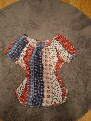Mönstrad blus med kort ärm - Säljer en luftig blus med kort ärm och rund hals. Toppen har ett färgglatt mönster i rött, blått, vitt och beige med inslag av blommor och ränder. Perfekt för dig som gillar boho-vibbar och vill ha något bekvämt och snyggt till sommaren.