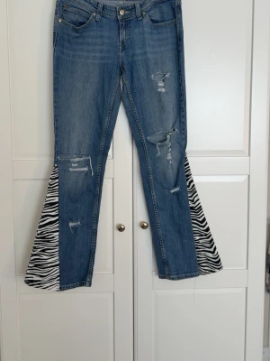 Blå utsvängda jeans med zebramönster - Unika blå jeans med slitningar och utsvängda ben där insidan har zebramönstrade tygpaneler. Jeansen har klassisk femficksmodell och knappgylf. Perfekta för dig som vill sticka ut med en cool och trendig look.