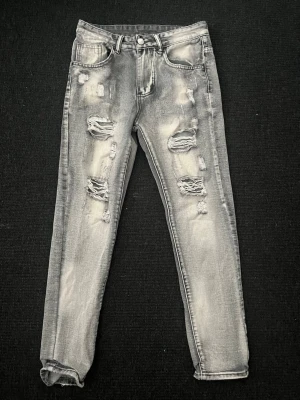 Grå slitna jeans med hål - Säljer ett par grå jeans med kraftiga slitningar och hål på framsidan. Jeansen har fem fickor, raka ben och en tvättad look som ger en cool och avslappnad stil. Perfekta för dig som gillar streetwear och trendiga detaljer.