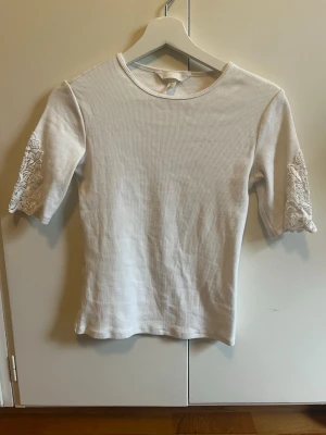 Vit ribbad topp med spetsdetaljer - Säljer en vit ribbad topp med halvlånga ärmar och fina spetsdetaljer på ärmarna. Toppen har en rund halsringning och sitter figurnära. Perfekt för dig som gillar en enkel men ändå söt stil.