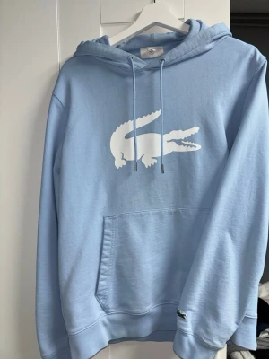 Ljusblå tracksuit från Lacoste - Snygg ljusblå tracksuit från Lacoste med stor vit krokodil-logga på bröstet. Hoodien har huva med dragsko, magficka och ribbade muddar. Tillverkad i mjukt material som känns skönt mot huden. Perfekt för en avslappnad och trendig look.
