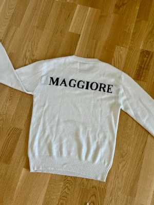 Vit stickad tröja med text - Vit stickad tröja med svart texten 'MAGGIORE' på ryggen. Tröjan har rund halsringning, långa ärmar och ribbade muddar vid ärmslut och nederkant. Enkel och stilren design som passar till mycket. Storleken är L(ungdom) motsvarar 170 eller S i herr