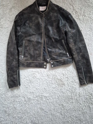  Skinnjacka från Nelly - Cool brun skinnjacka från Nelly med asymmetrisk dragkedja, två snedställda fickor med dragkedja och detaljerade sömmar. Jackan har en kortare modell och dragkedjor vid ärmsluten för extra edge.