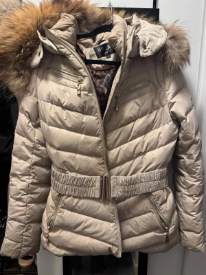 Beige dunjacka med pälshuva Hollies - Snygg beige dunjacka från Hollies med avtagbar huva och fejkpäls. Jackan har bälte i midjan, dragkedjor på fickor och bröst, samt leopardmönstrat innerfoder. Perfekt för kalla dagar och ger en riktigt trendig look. Det är svaga smuts fläckar här och där så en kemtvätt är att rekommendera. Säljer den för den var för liten när jag köpte den i våras på annan sida. Skriver mycket bra skick för det är den om man bortser från smutsen som är. 