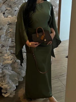Olivgrön långklänning med vida ärmar - Elegant olivgrön långklänning med vida, flödande ärmar och rund halsringning. Klänningen har en enkel, stilren design och är tillverkad i ett mjukt, slätt tyg som ger ett exklusivt intryck. Perfekt för dig som vill ha en unik och trendig look. Man kan knyta åt den bakifrån