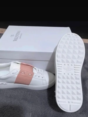 Valentino vita sneakers med rosa detalj - Stilrena vita sneakers från Valentino Garavani med bred rosa rem över mitten. Skorna har klassisk låg profil, vita snören och diskret logga på sulan. Tillverkade i slätt läder med gummisula för extra komfort och grepp.