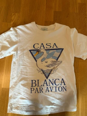 Vit t-shirt med svanmotiv - Säljer en vit t-shirt med tryck av en svan och texten 'Casa Blanca Par Avion' på framsidan. T-shirten har klassisk passform och korta ärmar. Materialet känns som bomull och trycket har blå och ljusblå detaljer. Skicket på den är jättebra förutom att trycket har spruckit lite därför priset e lågt men inget man märker på avstånd.
