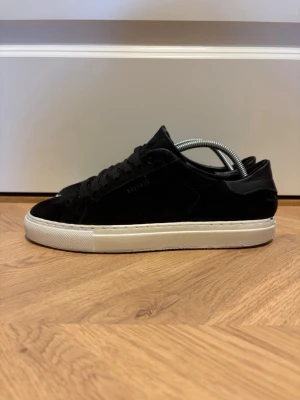 Axel Arigato Skor  - Säljer ett par svarta Arigato‑sneakers i mocka, storlek 38. Skorna är i mycket bra skick och fungerar helt som de ska. Endast en liten detalj: Arigato‑loggan på ena skon har nästan nötts bort, men det syns knappt vid användning.