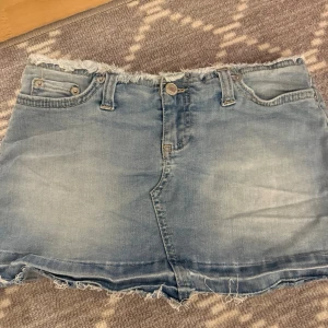 Ljusblå jeanskjol med fransig kant - Säljer en ljusblå jeanskjol med fransig, rå kant både upptill och nedtill. Kjolen har klassiska fem fickor, bälteshällor och knappgylf. Perfekt för dig som gillar en avslappnad och trendig stil.
