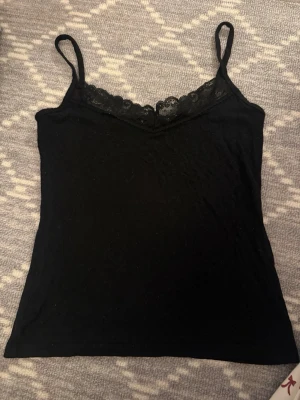 Svart linnetopp med spetsdetalj - Snygg svart linnetopp med tunna axelband och spetsdetalj vid urringningen. Perfekt att styla lager på lager eller bära som den är. Enkel och stilren design som passar till mycket.