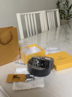 Svart Burberry bälte med silverdetalj - Snyggt svart bälte från Burberry med klassiskt rutigt mönster och stor silvrig B-spänne framtill. Bältet är gjort i skinn och kommer med originalpåse, box och certifikat. Perfekt accessoar för att lyfta din outfit.