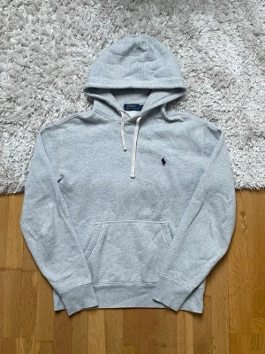 Grå hoodie från Polo Ralph Lauren - Snygg ljusgrå hoodie från Polo Ralph Lauren med klassisk huva och vita dragsnören. Har en broderad mörkblå logga på bröstet och en stor magficka. Tillverkad i mjukt bomullsmaterial som känns skönt mot huden. Perfekt för en avslappnad och stilren look.