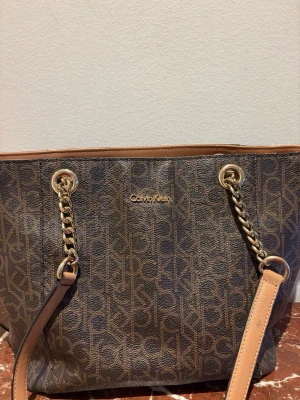 Brun handväska från Calvin Klein - Välj gärna instabox, kan skicka mycket snabbare då. Snygg brun handväska från Calvin Klein med guldiga kedjedetaljer och logomönster över hela väskan