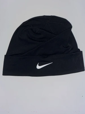 Svart mössa från Nike - Snygg svart mössa från Nike med klassisk vit swoosh-logga framtill. Tillverkad i mjukt och stretchigt material som sitter skönt på huvudet. Perfekt accessoar för kalla dagar och enkel att matcha med olika outfits.