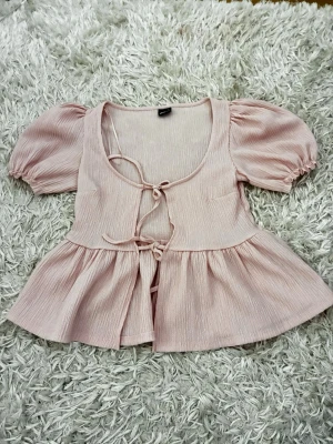 Ljusrosa blus med puffärm och knyt - Supersöt ljusrosa blus med puffärmar och två knyt framtill. Blusen har en lätt rynkad, volangig nederdel och är tillverkad i ett tunt, mjukt tyg som ger en romantisk vibe. Perfekt för dig som gillar feminina detaljer och en luftig känsla. Aldrig använd 
