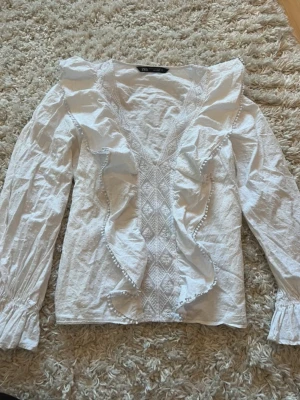 Vit blus med volanger från Zara - Supersöt vit blus från Zara med långa ärmar, djup v-ringning och dekorativa volanger framtill. Blusen har fina spetsdetaljer och smock vid ärmsluten. Perfekt för dig som gillar romantisk och trendig stil.