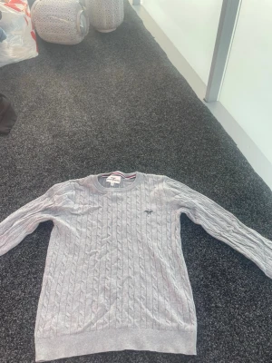 Ljusgrå kabelstickad tröja från hampton republic - Snygg ljusgrå kabelstickad tröja med rund halsringning och diskret broderad logga på bröstet. Tröjan har långa ärmar och ribbade muddar vid ärmslut och nederkant. Perfekt för lager på lager och enkel att matcha.