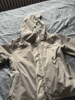 Beige vindjacka Peak Performance M - Snygg beige vindjacka från Peak Performance med huva och dragkedja framtill. Två fickor med dragkedja och diskret logga på bröstet. Tillverkad i lätt och tåligt material, perfekt för blåsiga dagar.