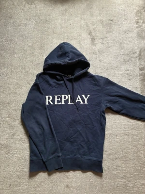 Replay hoodie - Snygg mörkblå hoodie från Replay med stor vit logga över bröstet. Klassisk modell med huva och dragsnören, ribbade muddar vid ärmslut och nederkant. Perfekt för en avslappnad och trendig look.