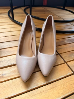 Beige klassiska pumps med spetsig tå - Säljer ett par stilrena pumps i beige med spetsig tå och låg klack. Skorna har en slät yta i syntetmaterial och svart sula. Perfekta för dig som gillar en enkel men elegant look.