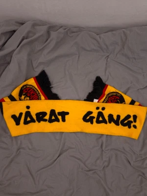 Luleå Hockey halsduk gul och svart - Gul Luleå halsduk med svart text 'VÅRAT GÄNG!' och Luleå Hockeys logga i svart och rött. Svarta och röda ränder samt fransar i ändarna. Perfekt för att visa ditt stöd på matchen eller i vardagen.